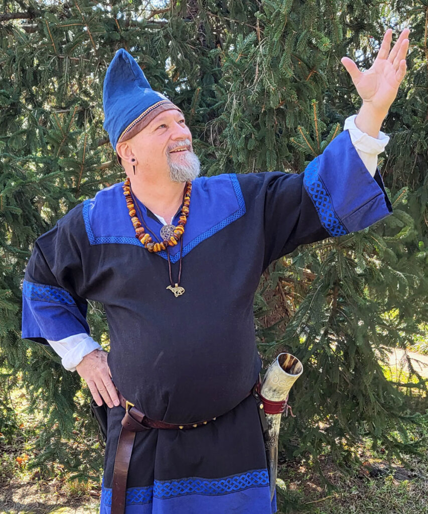 Man wearing a blue and black Viking Age linen tunic, belt, and hat, representing early medieval Scandinavian dress.